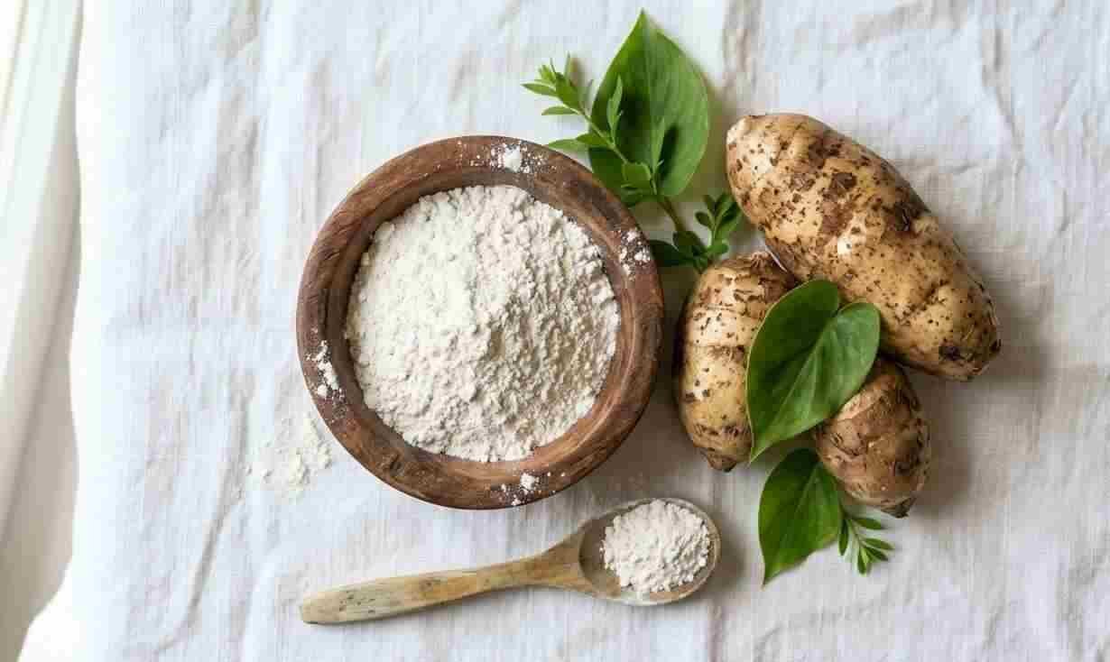 White arrowroot powder in a wooden bowl with fresh arrowroot tubers and green leaves on a rustic kitchen counter